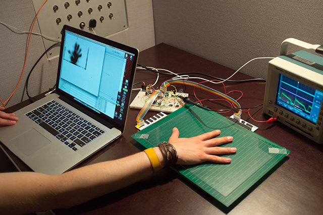 The sensor plate is connected to a breadboard, a macbook and an oscilloscope. The display of the macbook shows the outlines of the hand which rests on the sensor plate.